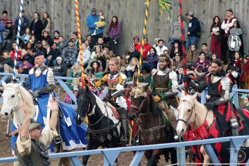 A group of knights on horseback in armor, preparing for a jousting event, with a crowd watching in the background.