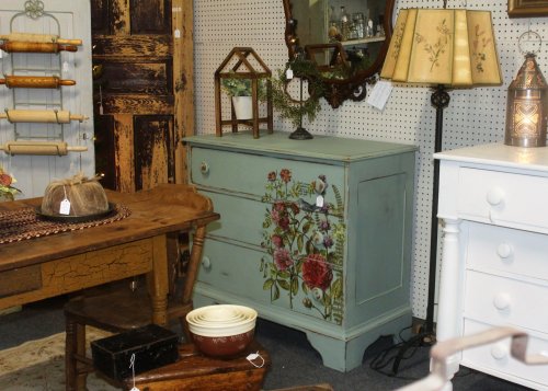 A vintage room featuring a floral-painted dresser, wooden table, and various decorative items in a cozy setting.