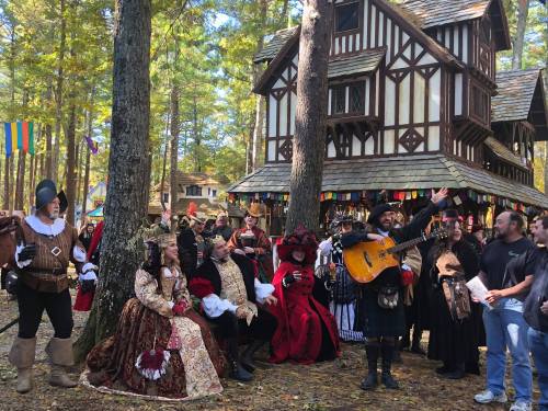 A lively gathering of people in medieval costumes, with a musician playing guitar in front of a rustic building in a forest.