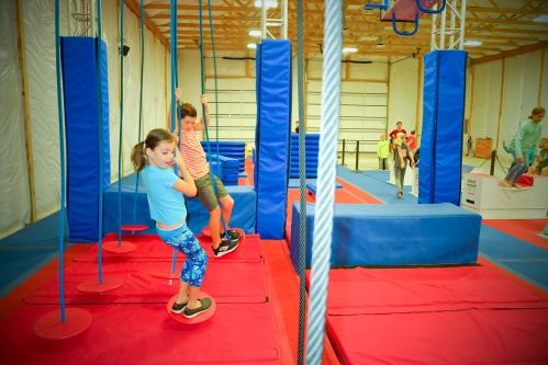 Two children play on gym equipment in a colorful indoor obstacle course, with others in the background.