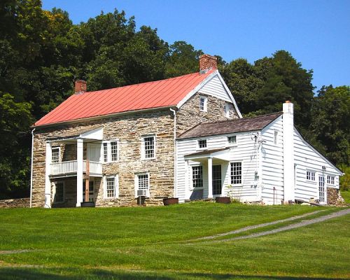 Historic stone and wooden house with a red roof, surrounded by green grass and trees under a clear blue sky.