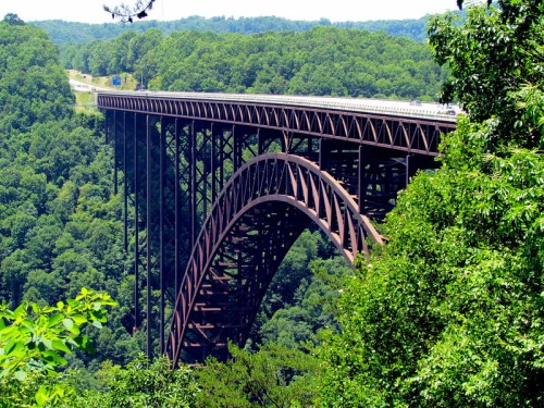 A large, arched metal bridge spans a lush green valley, surrounded by dense trees and a clear blue sky.