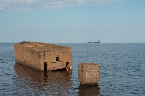 A large concrete structure partially submerged in calm water, with a ship in the distance under a clear blue sky.
