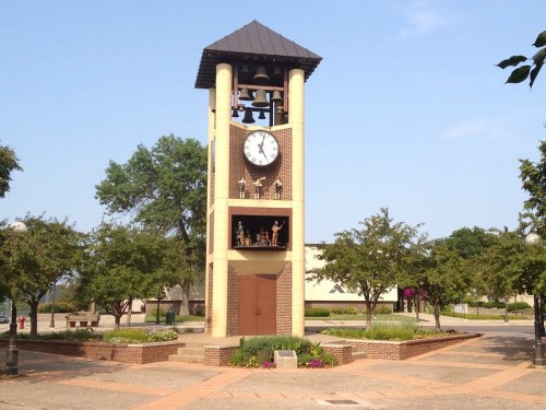 A tall clock tower with bells, surrounded by trees and greenery in a public square on a clear day.