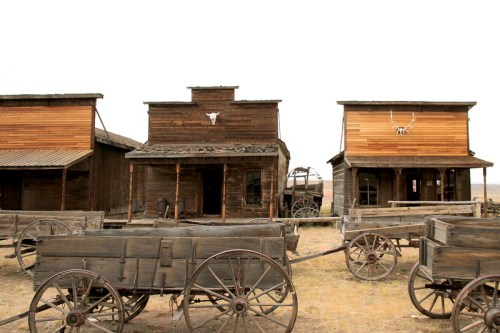 Old Western town with wooden buildings and vintage wagons in a dusty landscape.