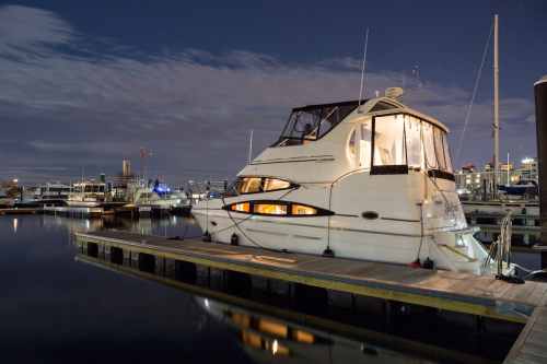A lit yacht docked at a marina at night, reflecting in calm water with city lights in the background.