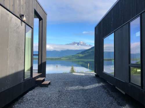 Two modern cabins frame a scenic view of a lake and mountains under a clear blue sky.