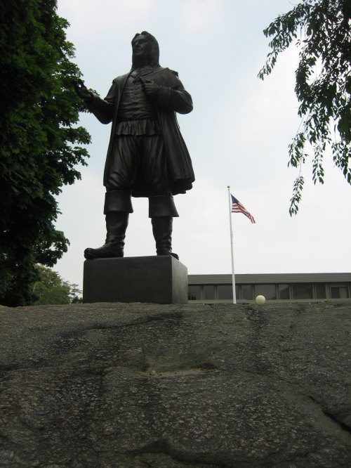 Statue of a historical figure on a rocky base, with an American flag in the background and trees nearby.