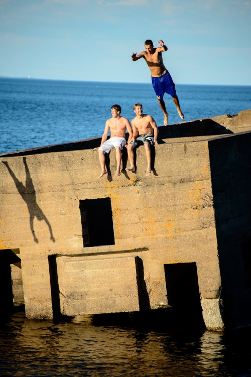Three young men on a concrete structure by the water; one is jumping off while the others sit nearby.