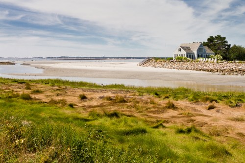 A serene beach scene featuring a house near the shore, with grassy areas and a calm, sandy landscape under a blue sky.