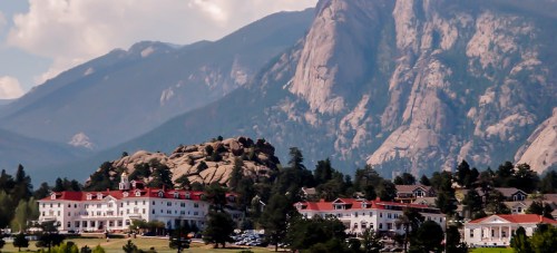 A large white hotel with red roofs sits at the base of rocky mountains under a blue sky with scattered clouds.