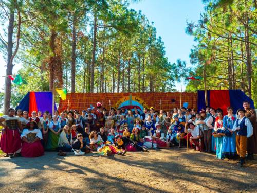 A large group of people in colorful costumes poses together in a forest setting, with festive decorations in the background.