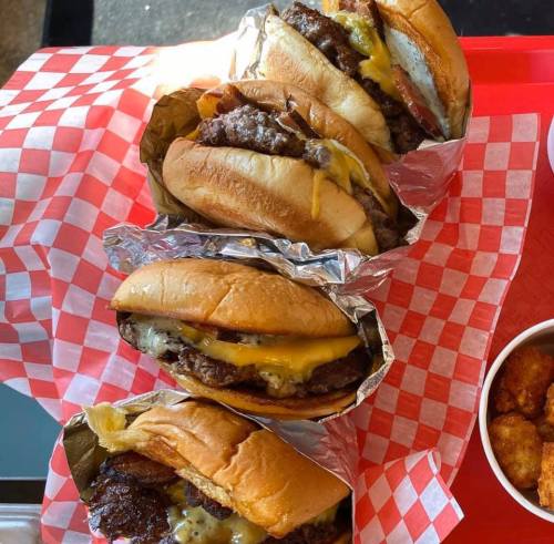 Three delicious burgers wrapped in foil, topped with cheese and served on checkered paper, alongside a bowl of tater tots.