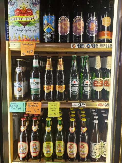 A display of various beer bottles and soft drinks on shelves, with price tags visible for each item.