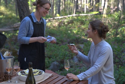 Two women enjoy a meal outdoors in a forest, with wine glasses and a platter of food on a wooden table.