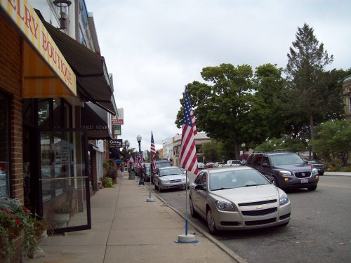 A street scene featuring parked cars, American flags, and shops under a cloudy sky.