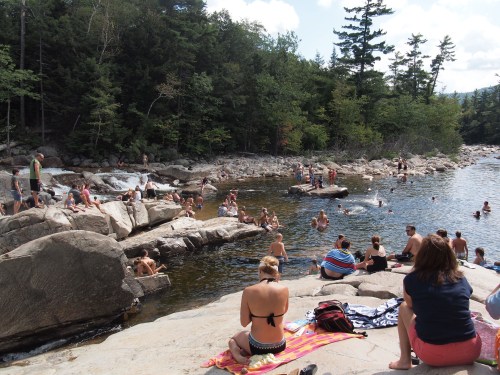 A crowded riverbank scene with people swimming and sunbathing on rocks surrounded by trees and water.