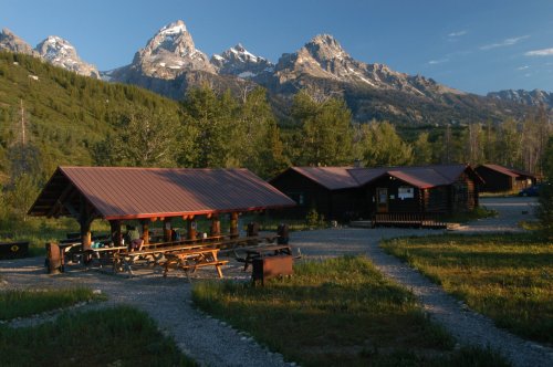 A rustic cabin area surrounded by lush greenery and towering mountains under a clear blue sky.