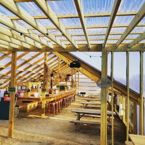 Interior of a rustic greenhouse with wooden beams, long tables, and plants, creating a cozy dining atmosphere.