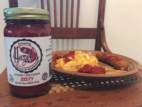A jar of Hazel's Zesty gourmet chili sauce next to a plate of scrambled eggs and sausage, with sauce drizzled on top.