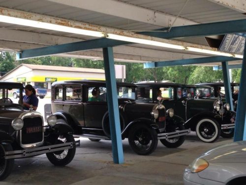 A row of vintage 1920s cars parked under a covered area, with people nearby.