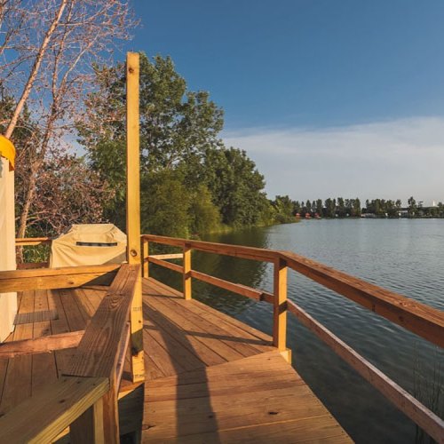 A wooden deck overlooking a calm lake, surrounded by trees and a clear blue sky.