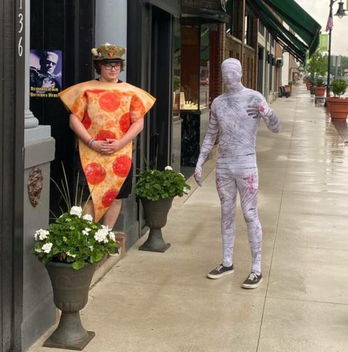 A person in a pizza costume stands by a building, while another in a mummy outfit poses nearby on a rainy street.