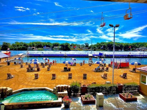 A sunny beach scene with chairs, umbrellas, and a lake in the background, surrounded by trees and blue skies.