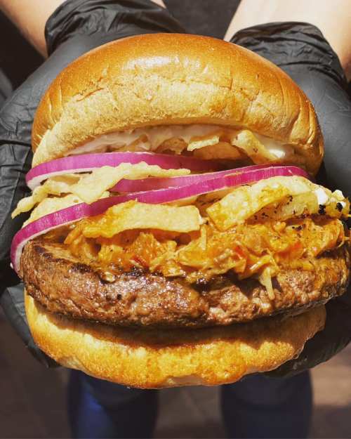 A close-up of a juicy burger with a beef patty, topped with shredded cabbage, onions, and crispy toppings in a bun.