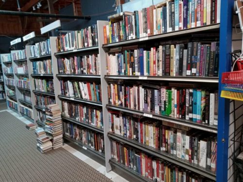 Bookshelves filled with various books in a library, with a stack of books on the floor to the left.