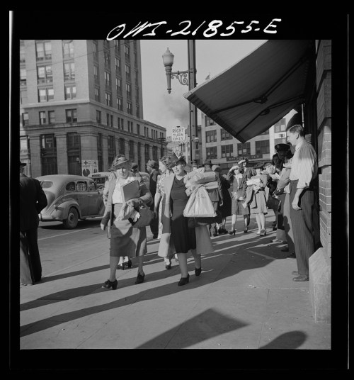 A busy street scene with women in hats and dresses walking, some carrying children, and a man standing by a building.
