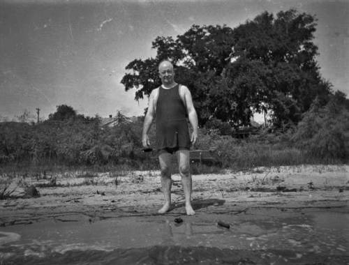 A man in a vintage swimsuit stands on a beach, with trees and a shoreline in the background.