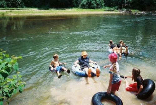 A group of children and adults enjoying a sunny day by the river, floating on inner tubes and playing in the water.