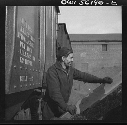 A man leans out of a train car, smiling, with industrial buildings in the background.
