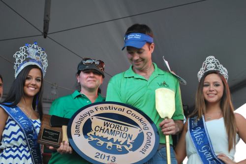 Three women in crowns and sashes stand beside a man holding a trophy and a cooking spatula at a festival event.