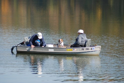 Two men in a small boat on a calm lake, accompanied by a dog, surrounded by autumn-colored trees.