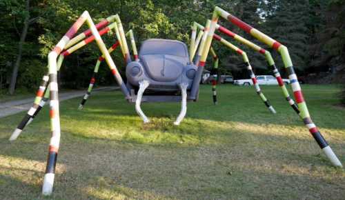 A large spider sculpture made from colorful pipes, with a car body as its body, set in a grassy area.