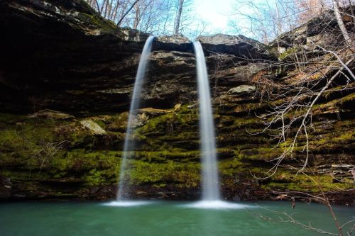 A serene waterfall cascading over rocky cliffs into a tranquil green pool, surrounded by trees and blue sky.