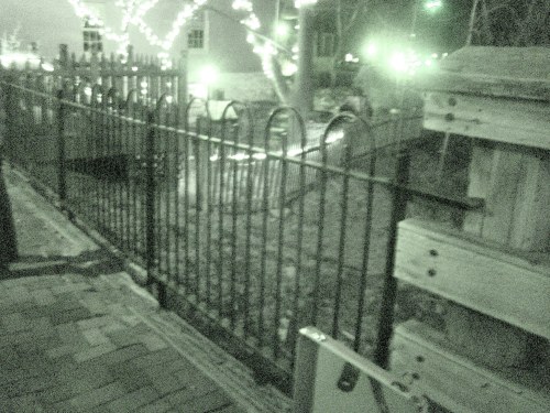 A dimly lit scene of a metal fence with decorative arches, surrounded by trees and soft lights at night.