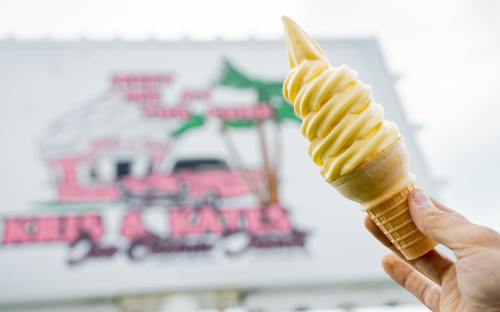 A hand holds a soft-serve ice cream cone in front of a colorful ice cream shop sign.