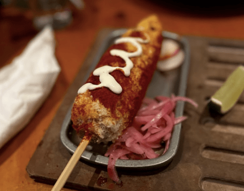 A grilled corn on a stick topped with mayonnaise and spices, served with pickled onions and a lime wedge.