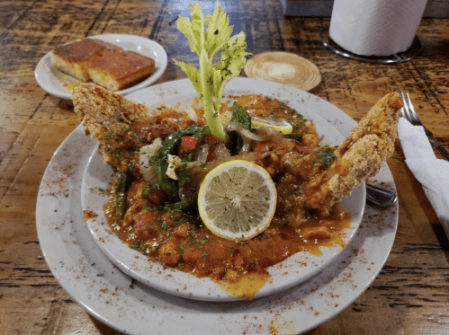 A plate of seafood stew garnished with lemon and celery, served with cornbread on the side.