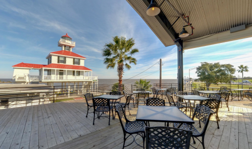 View of a waterfront restaurant patio with tables, palm trees, and a lighthouse in the background under a clear sky.