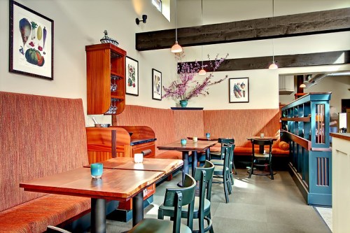 Cozy restaurant interior with wooden tables, green chairs, and decorative artwork on the walls. Soft lighting creates a warm ambiance.