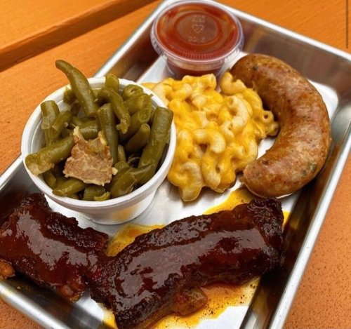 A metal tray with BBQ ribs, sausage, macaroni and cheese, and green beans, served with a side of sauce.