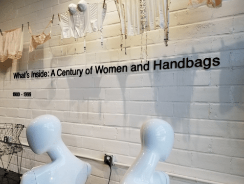 Exhibit title "What's Inside: A Century of Women and Handbags" displayed on a white wall with vintage clothing above.