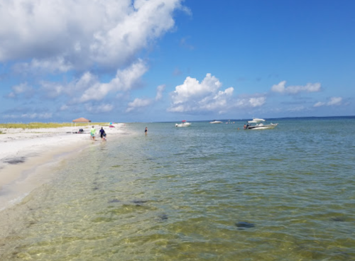 A serene beach scene with clear water, boats in the distance, and people walking along the shore under a blue sky.