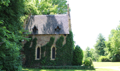 A small stone chapel covered in green ivy, surrounded by lush trees and bright sunlight.