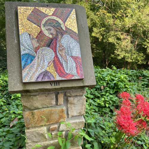 Mosaic of Jesus carrying the cross, mounted on a stone pillar, surrounded by greenery and red flowers.