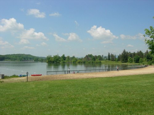 A serene lake scene with a sandy beach, green grass, trees, and fluffy clouds in a blue sky.